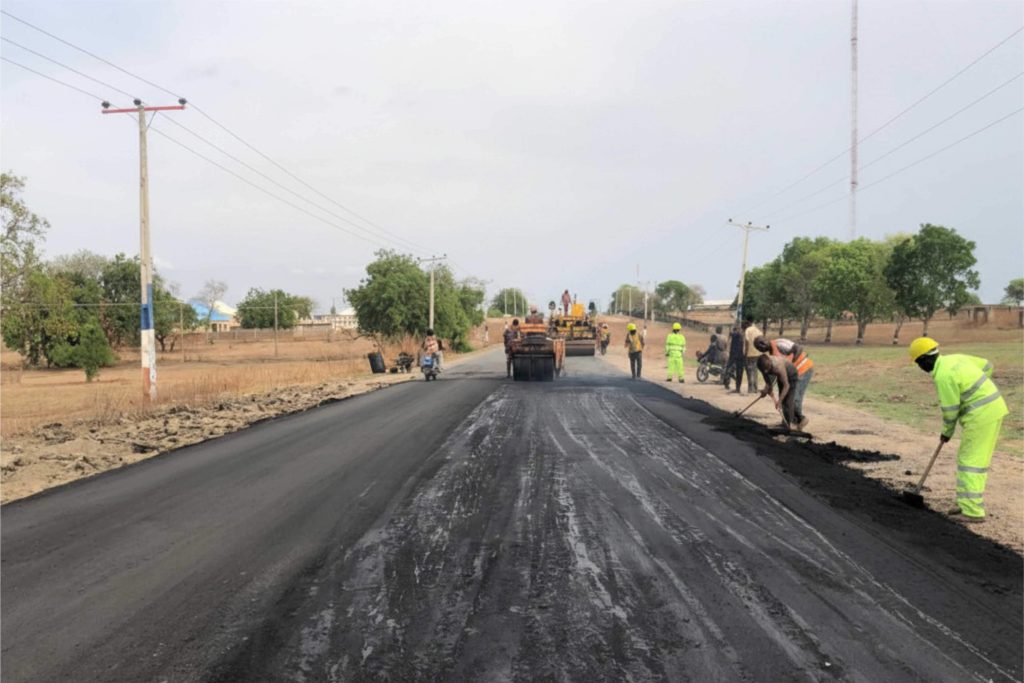 A group of construction workers wearing high-visibility lemon green jackets and safety helmets working on an asphalt road paving project with heavy machinery.